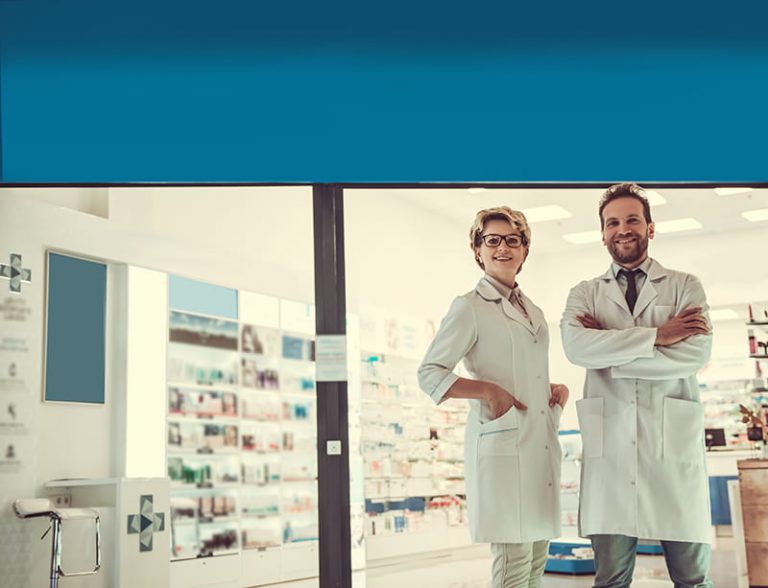 Imagen decorativa donde se ve a una mujer y un hombre farmacéuticos con bata blanca y sonrientes delante del escaparate de una farmacia, donde se aprecian a través del cristal estanterías con medicamentos